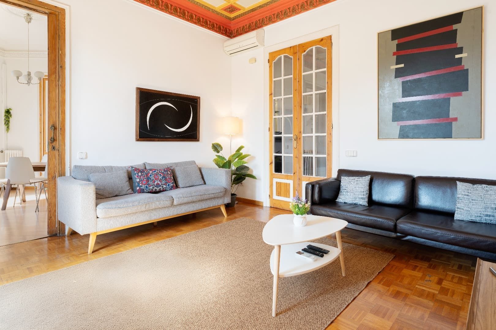 Sala de estar luminosa con sofá blanco, sillón de cuero negro, arte abstracto en la pared y mesa de centro redonda.