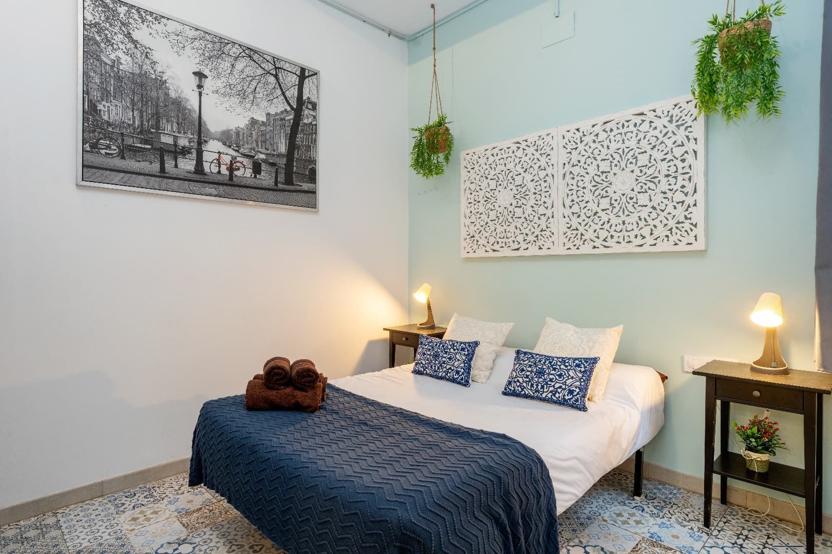 Habitación acogedora con ropa de cama azul, cuadros estampados en la pared, lámparas de noche y plantas colgantes.