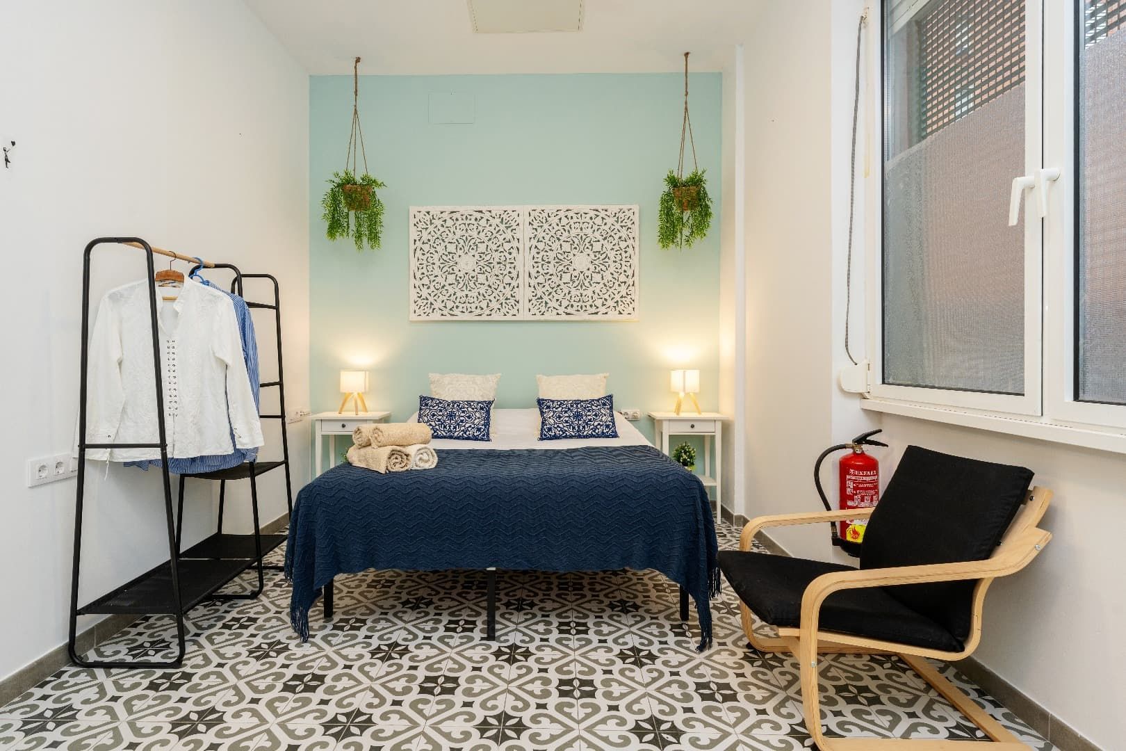 Habitación luminosa con colcha azul, suelo estampado, plantas colgantes y una silla negra junto a la ventana.