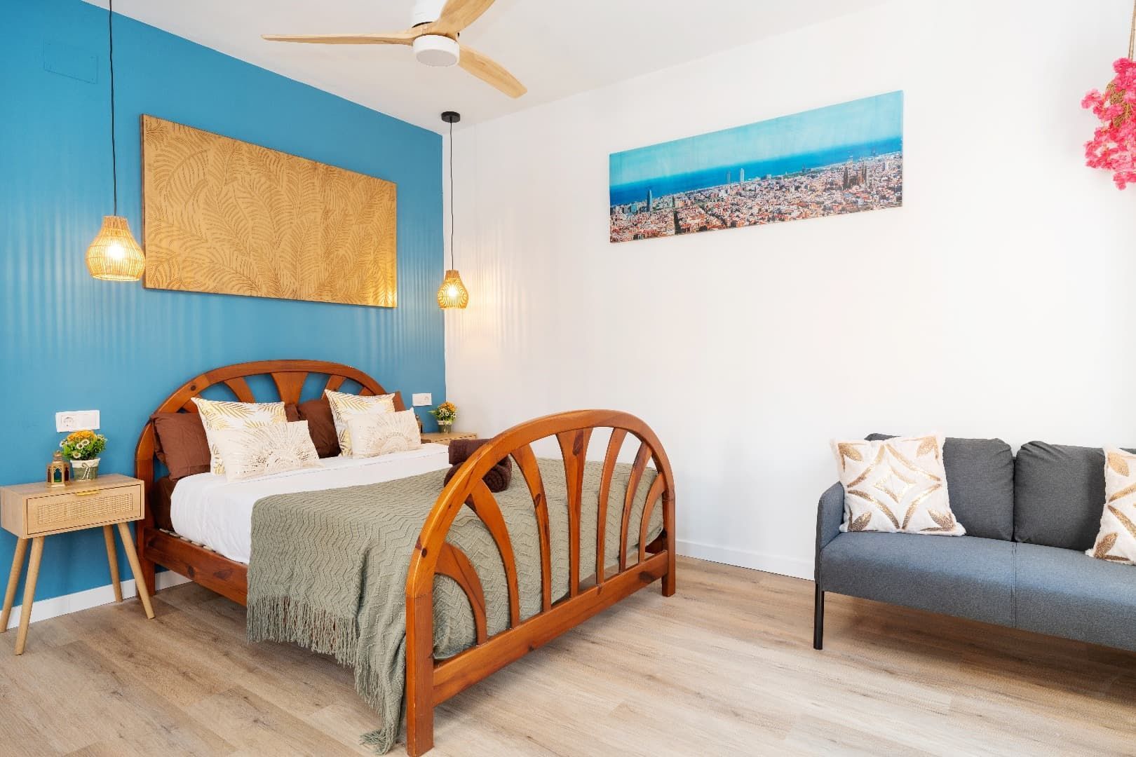 Dormitorio luminoso con pared decorativa color turquesa, cama de madera, sofá gris y cuadros con motivos playeros.