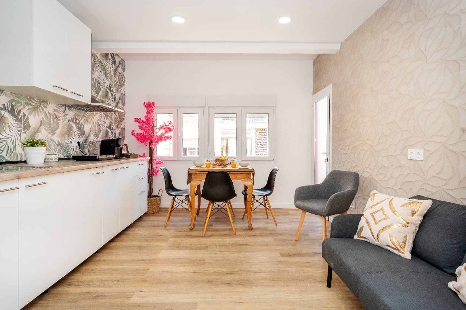 Luminoso salón-comedor moderno con suelo de madera, armarios de cocina blancos, sofá gris y mesa junto a puertas de cristal.