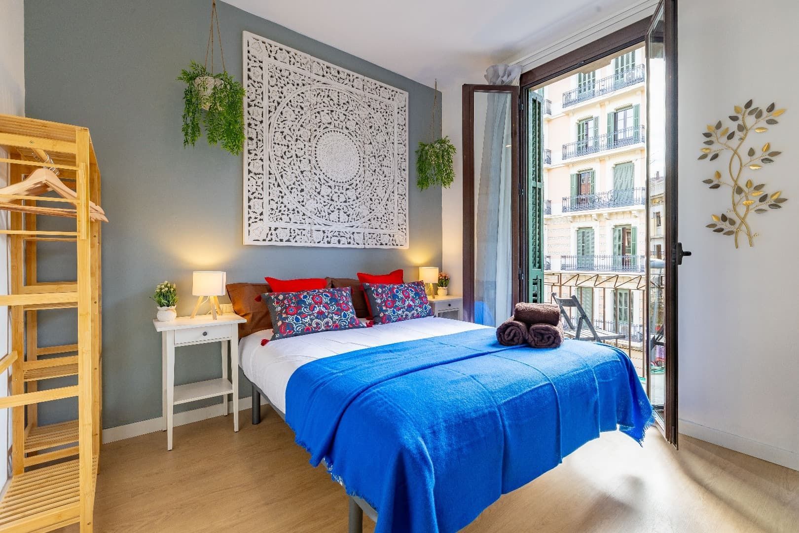 Habitación luminosa con colcha azul, cuadro decorativo con estampado, mesita de noche y gran ventanal con puerta al balcón.