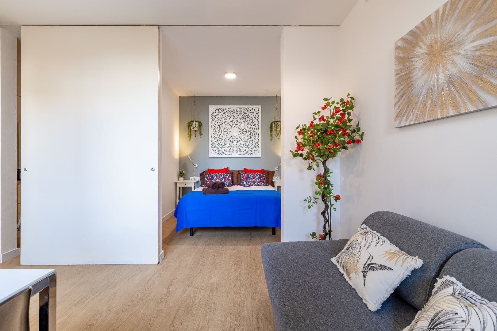 Habitación luminosa con colcha azul, sofá gris, planta de interior y arte abstracto en la pared.