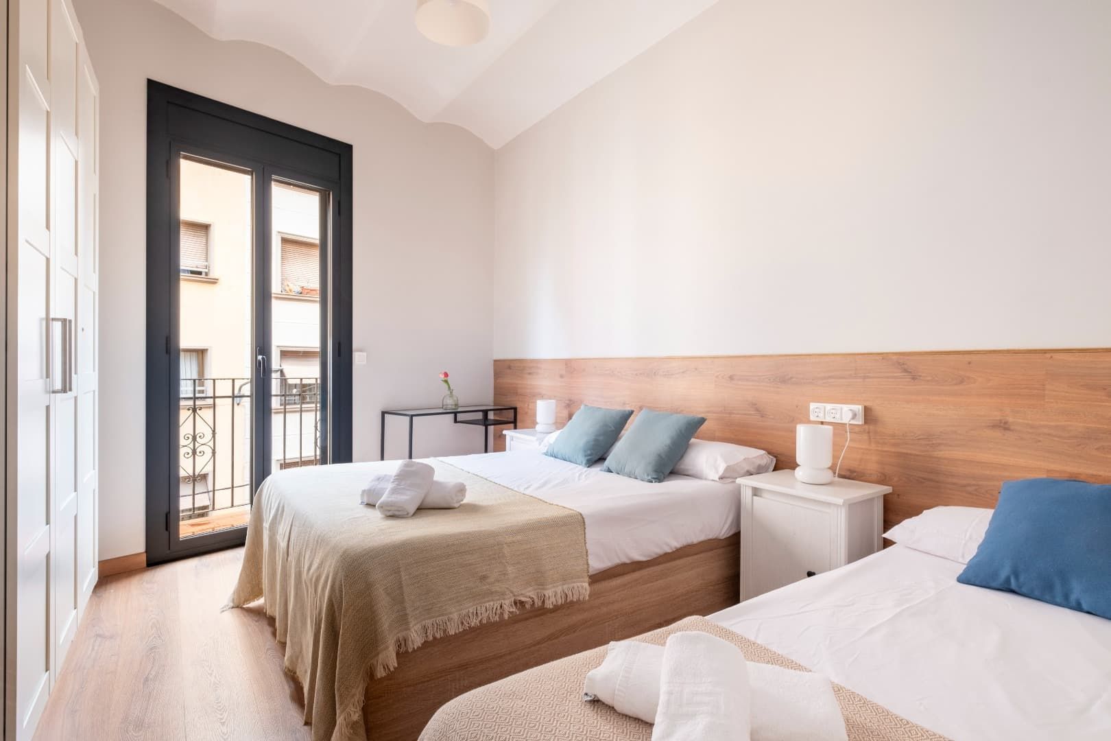 Habitación luminosa con dos camas individuales, cabecero de madera y puerta de balcón con contraventanas negras.