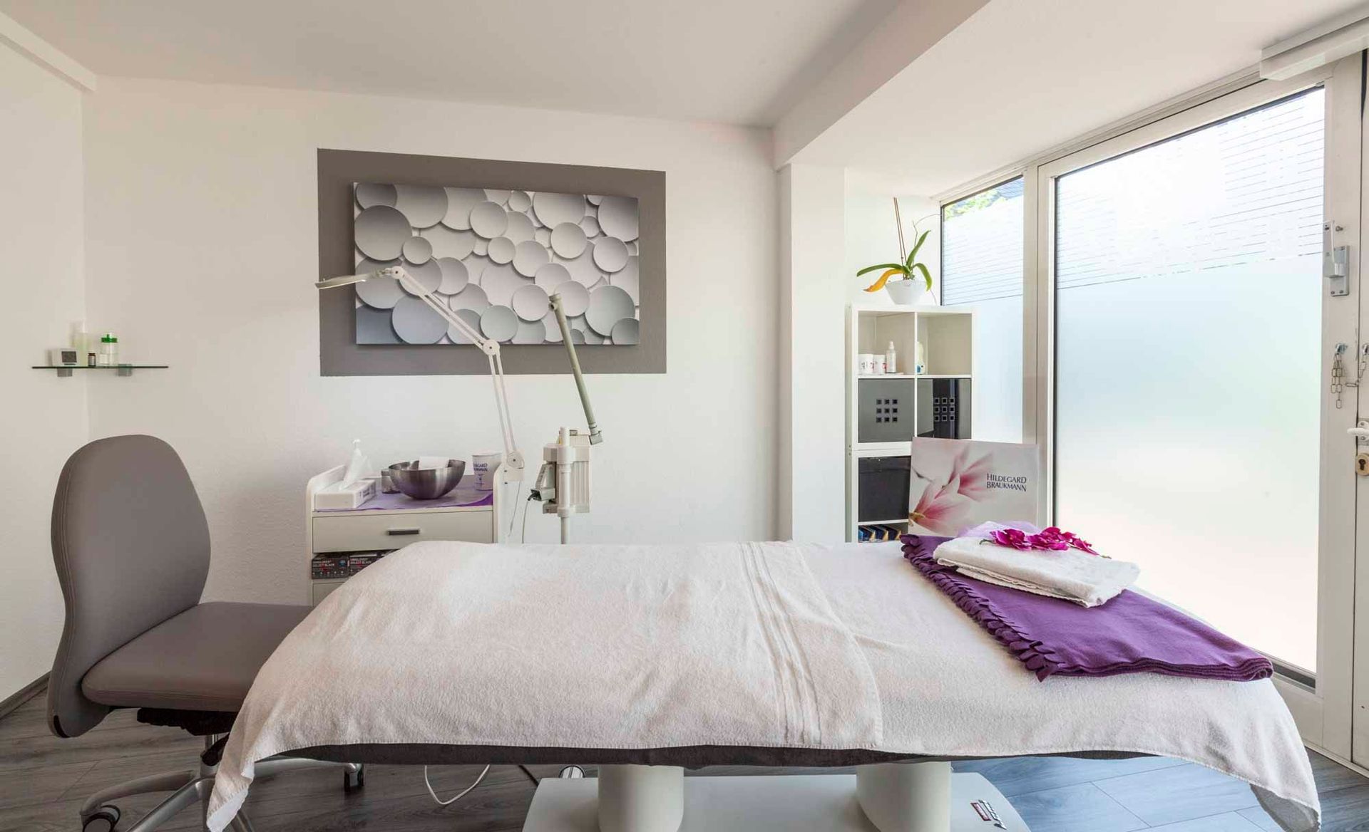 ein weißer Massagetisch mit einer lila decke und einem grauen Stuhl in der Beauty & Wellness Lounge