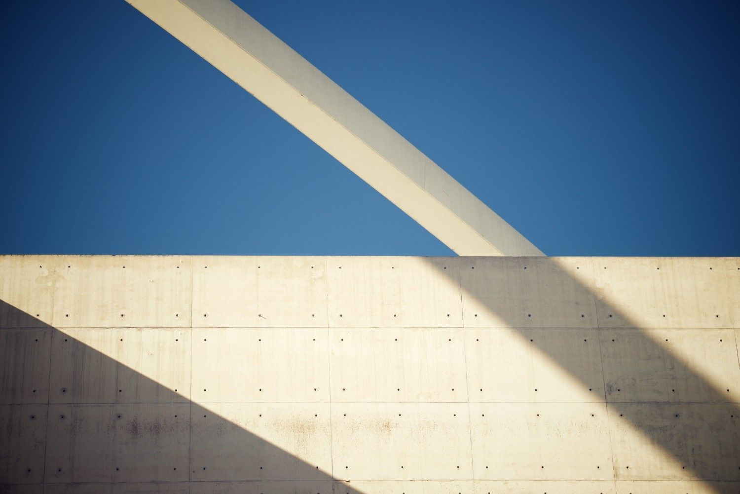 Cielo azul sobre un muro de hormigón beige con bandas de sombra diagonales proyectadas por una estructura superior.
