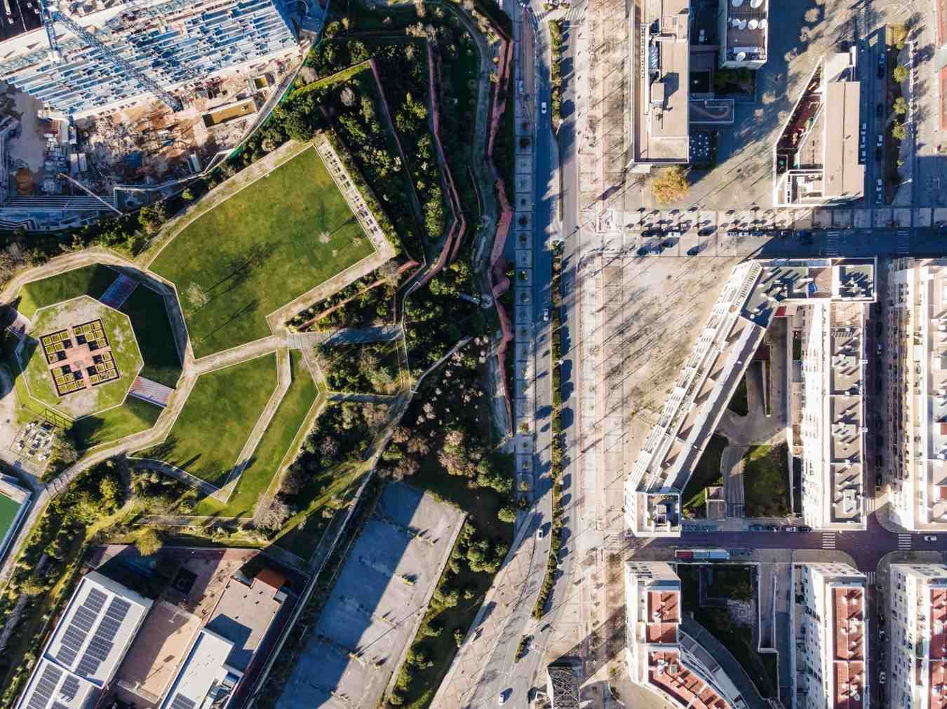 Vista aérea de un parque urbano junto a una carretera, rodeado de edificios y árboles frondosos.