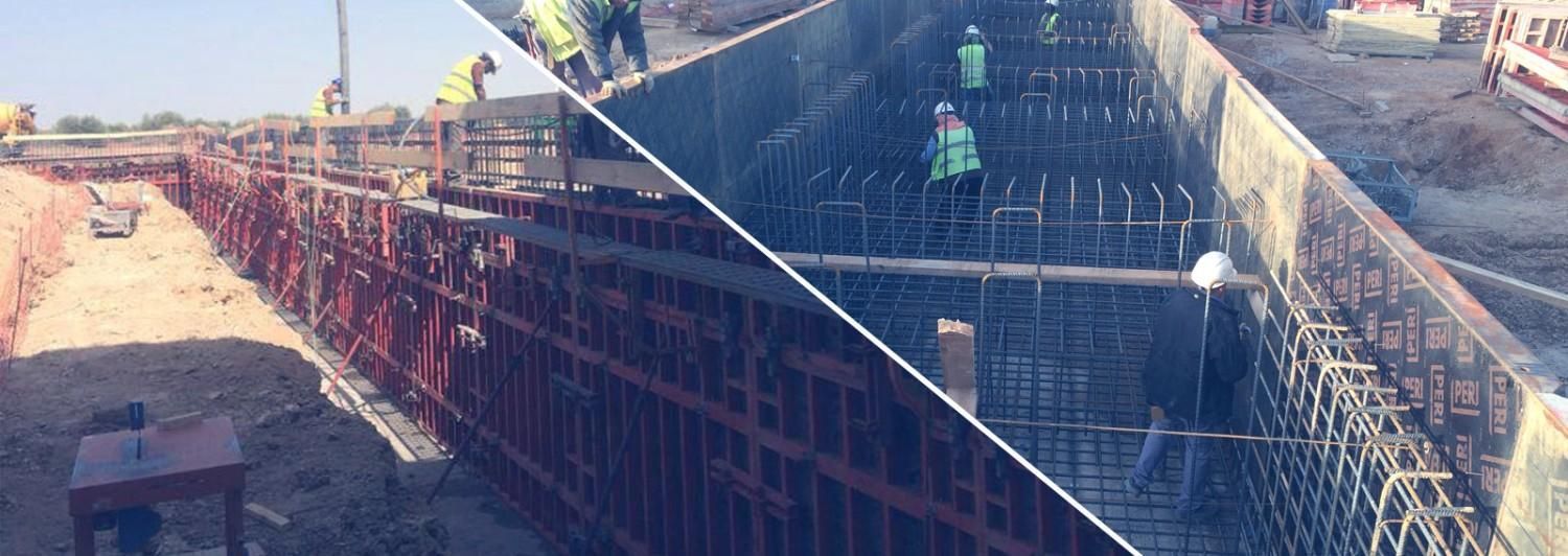 Obra en construcción con estructura de acero rojo y trabajadores en andamios cerca de un muro de hormigón inclinado.