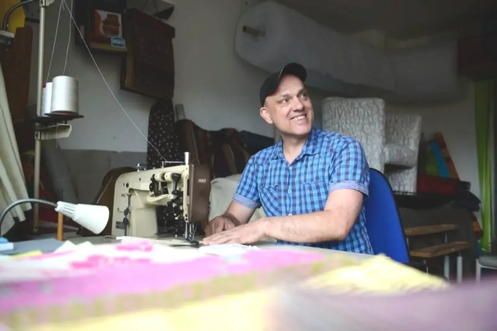 Ein Mann lächelt an einer Nähmaschine und arbeitet an einem Tisch. Werkstattumgebung mit Garnrollen und Stoffrollen.