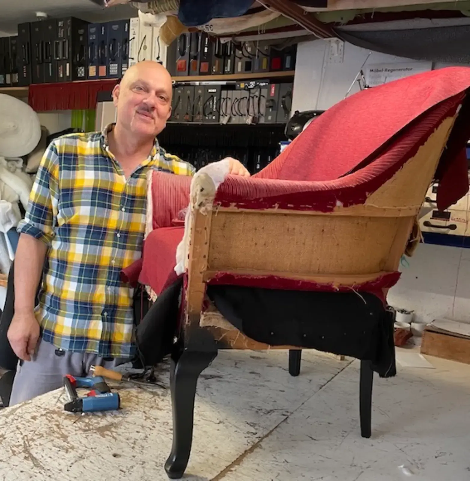 Ein Mann lächelt und steht neben einem teilweise neu bezogenen roten Stuhl in einer Werkstattumgebung.