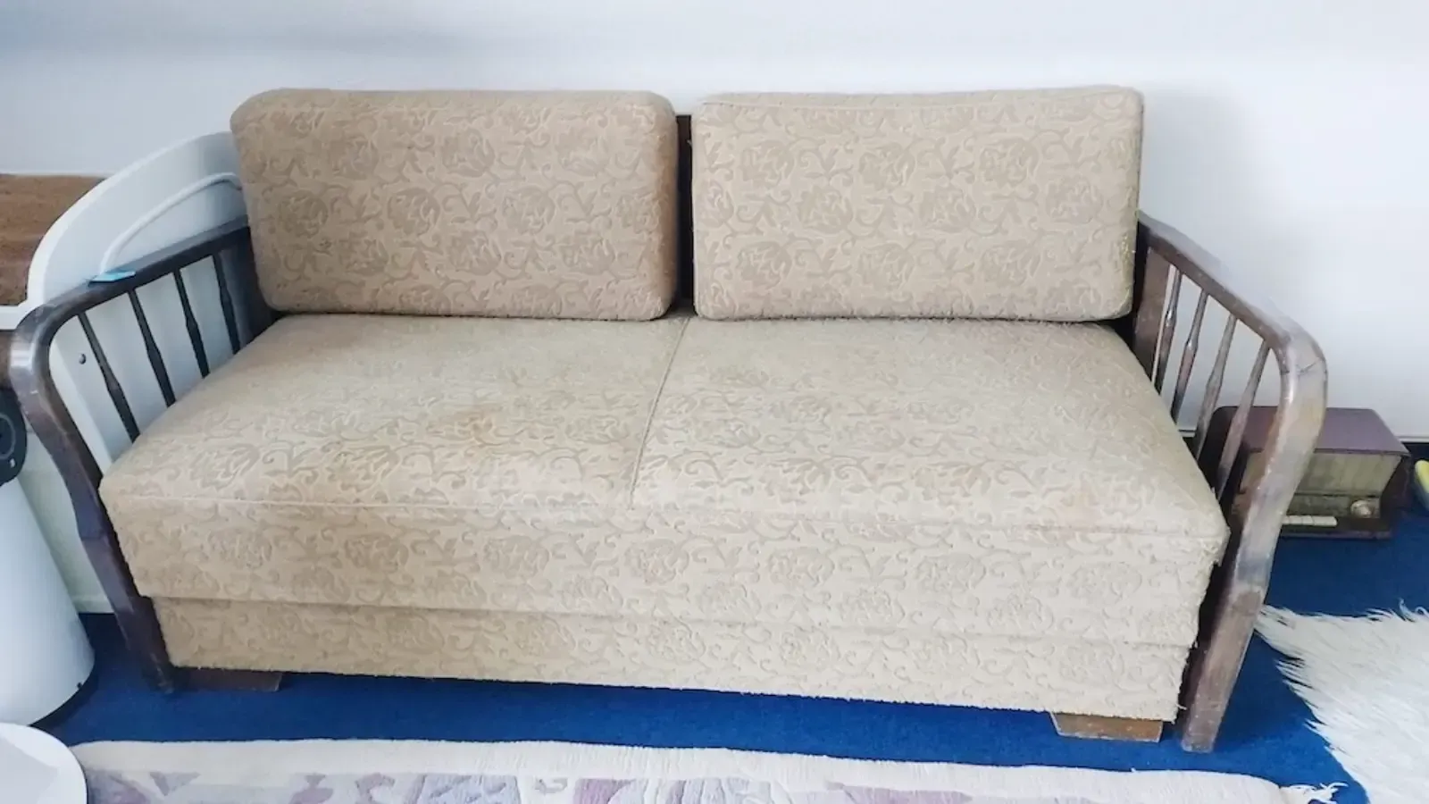 Beigefarbenes Zweisitzer-Sofa mit zwei Rückenkissen und Holzrahmen, auf einem blauen Teppich.