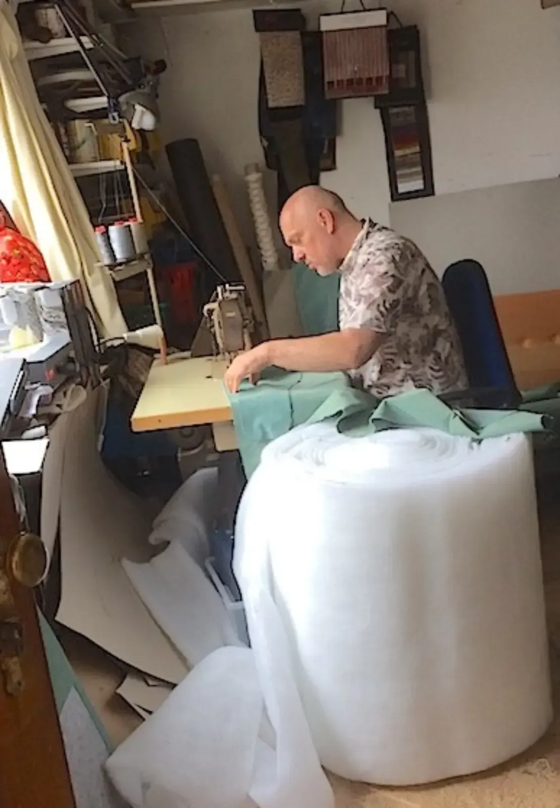 Ein Mann näht Stoff an einer Nähmaschine in einer Werkstatt, die mit Materialien gefüllt ist, darunter eine große Rolle weißer Schaumstoff.