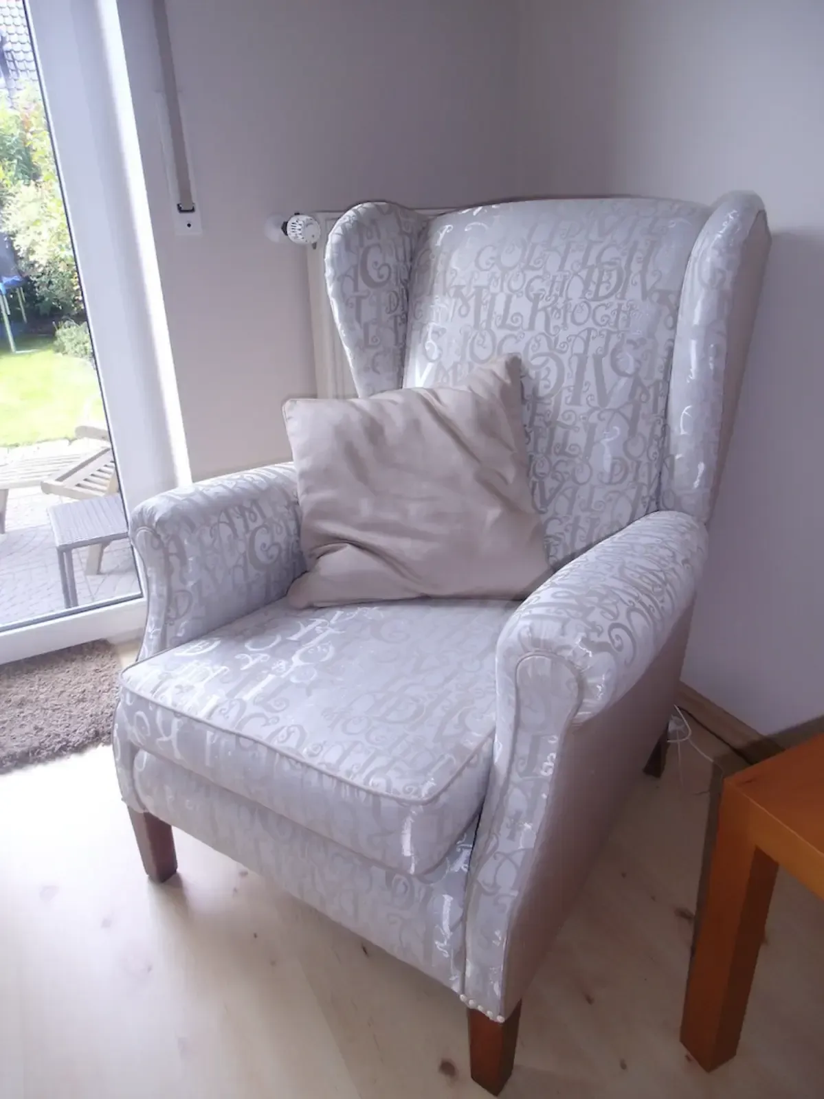 Ohrensessel mit gemustertem Stoffbezug, Kissen und Holzbeinen. In einem Zimmer mit Tür und Beistelltisch.