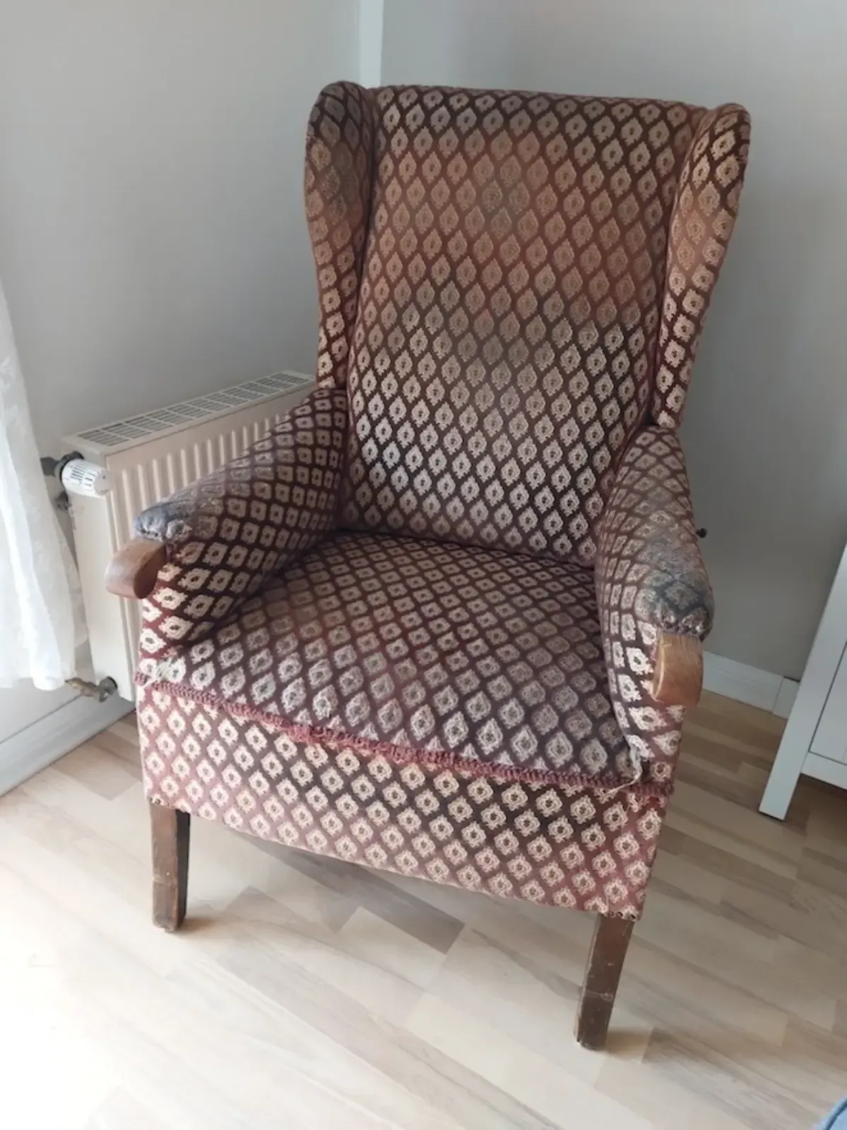 Ohrensessel mit braun gemustertem Bezug, Armlehnen und Beinen aus Holz, neben einem Heizkörper.