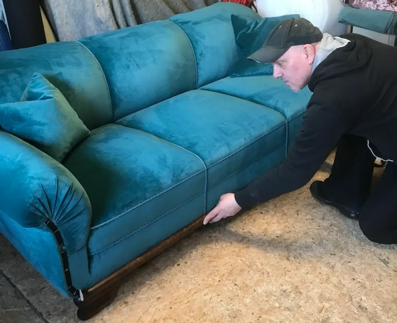 Ein Mann begutachtet in einer Werkstattumgebung das Gestell eines türkisfarbenen Samtsofas.