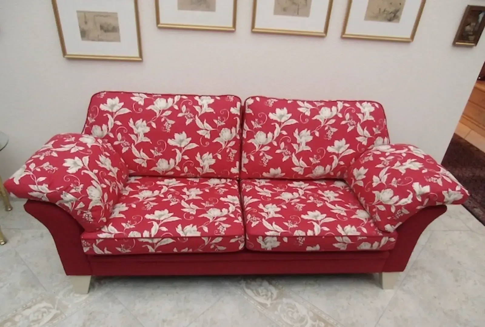 Ein rotes Sofa mit Blumenmuster und weißen Akzenten steht vor einer weißen Wand mit gerahmten Kunstwerken.