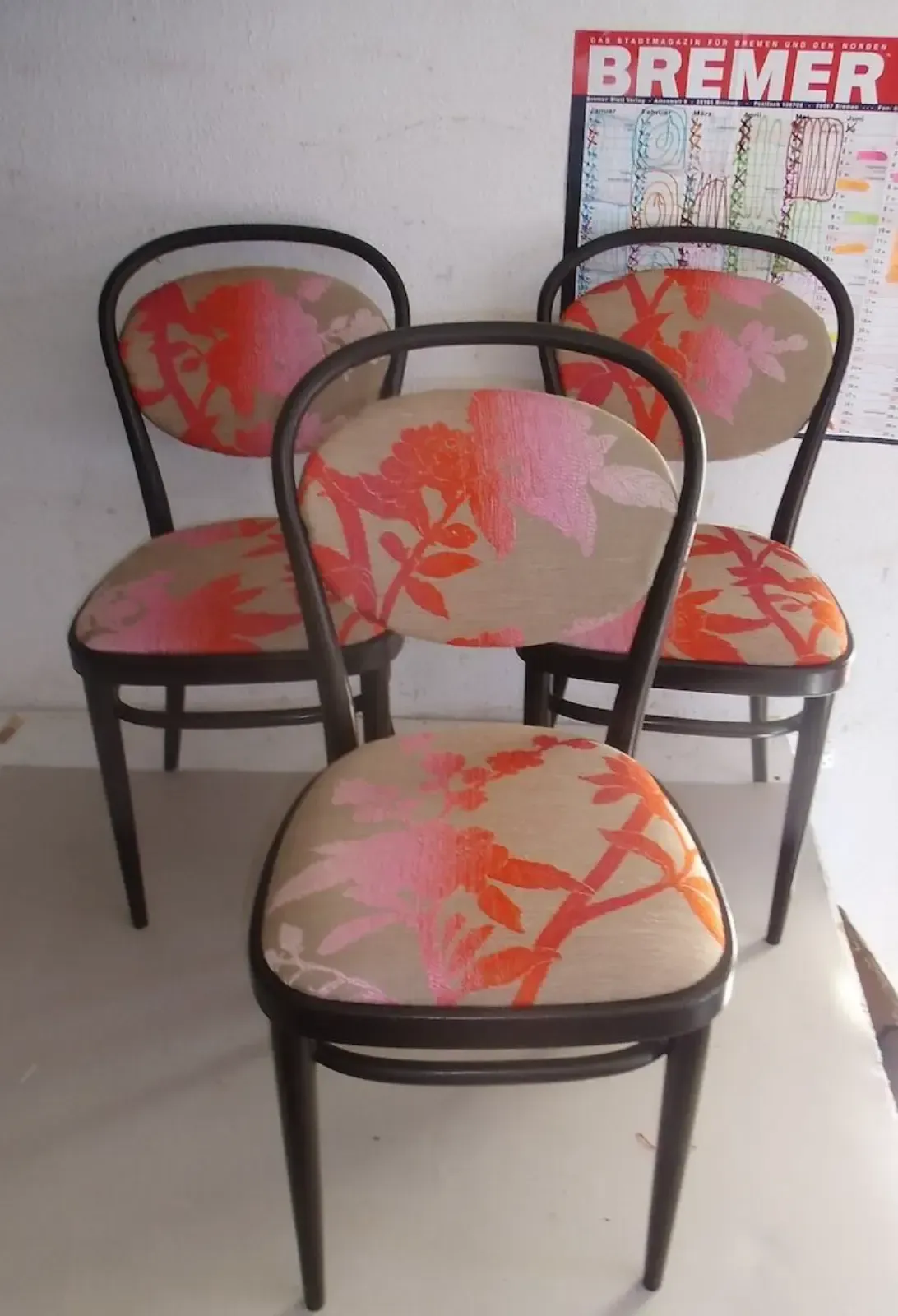 Vier Stühle mit braunem Gestell und rosa-orangefarbenem Blumenmuster in einem Zimmer mit einem Kalender.