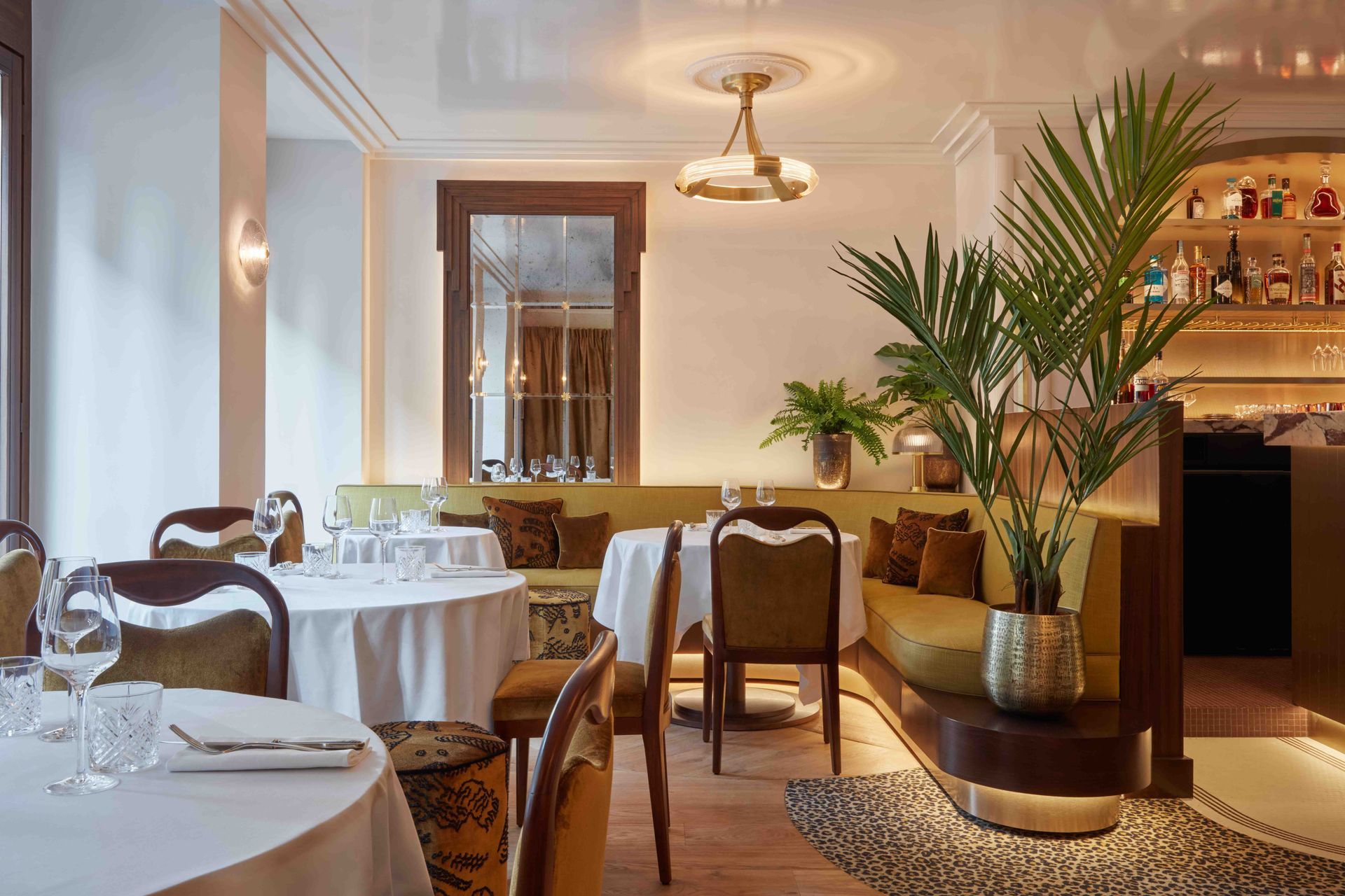 Élégante salle de restaurant avec des tables dressées, des nappes blanches et un palmier en pot près d’un bar.