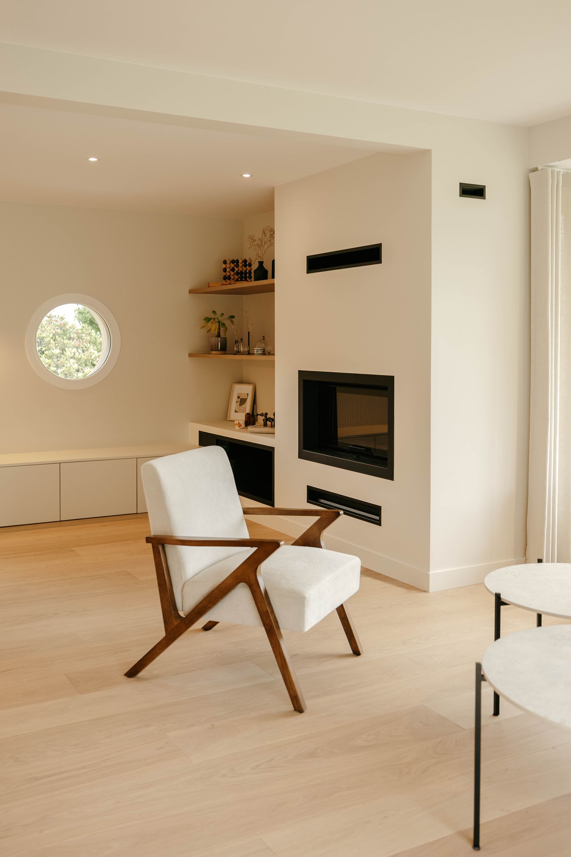 Salon minimaliste avec des murs beiges, un fauteuil blanc, une cheminée, une fenêtre ronde et un parquet clair