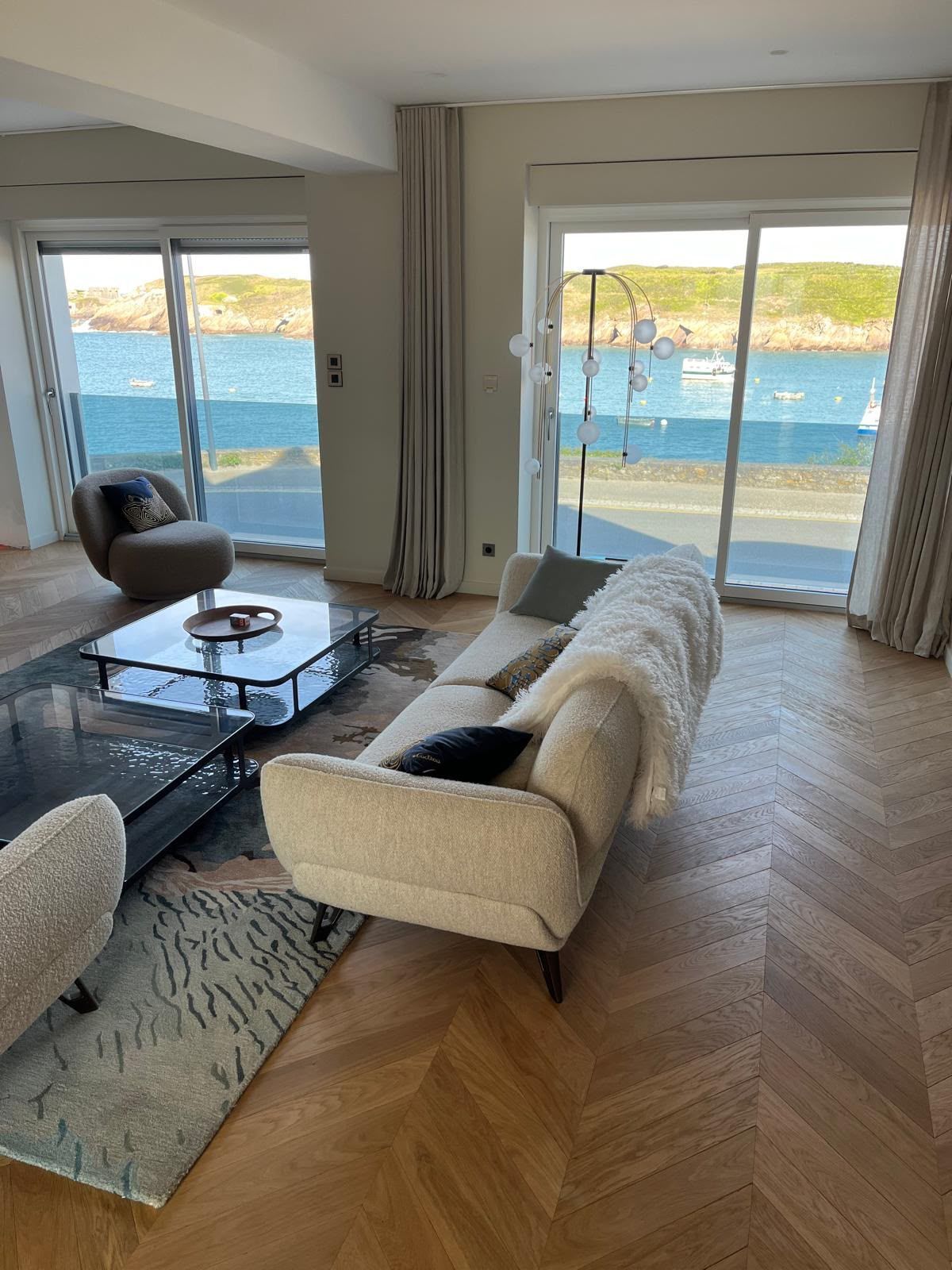Salon lumineux avec vue sur l’océan, canapé beige, table basse en verre et grandes fenêtres ouvertes sur la mer.