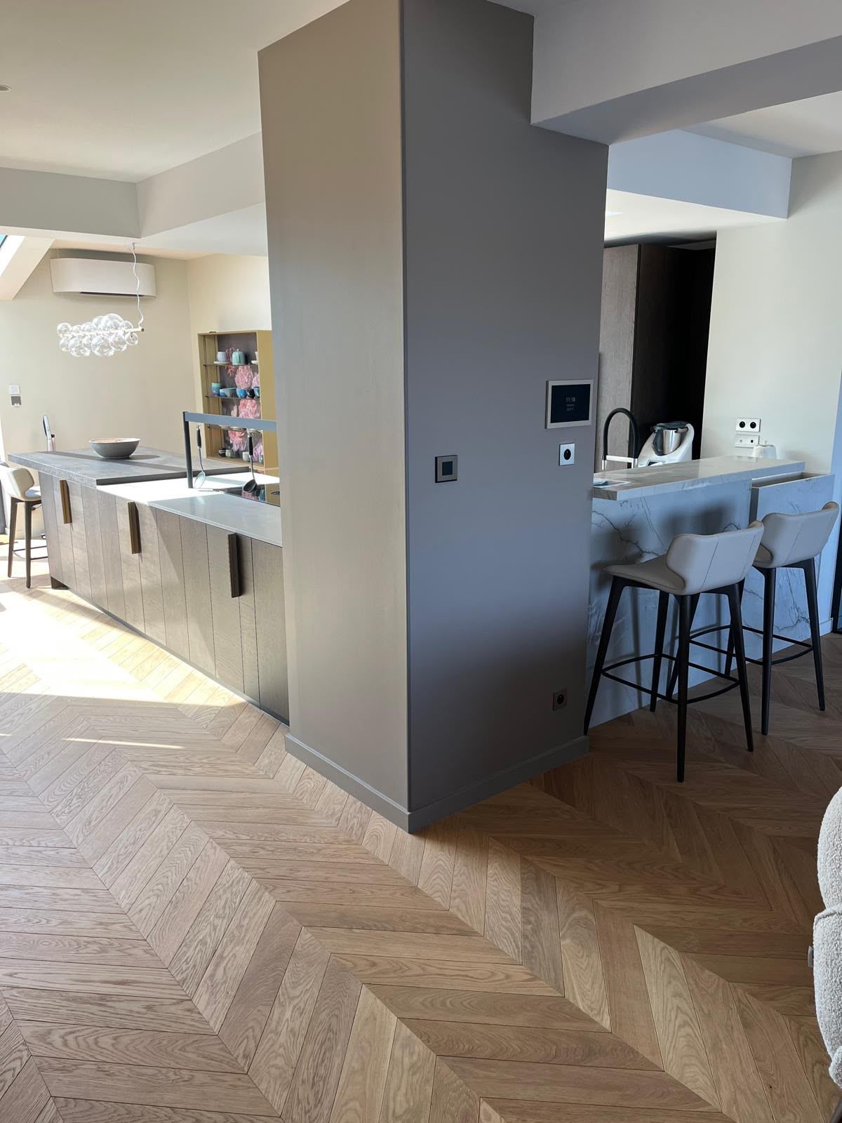 Pièce de vie ouverte avec un mur d’accent gris, des tabourets autour de l’îlot de cuisine et un parquet en point de Hongrie.