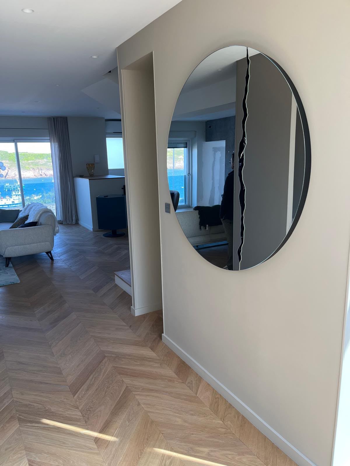 Couloir avec parquet en chevrons et miroir rond au mur, menant à un salon lumineux.