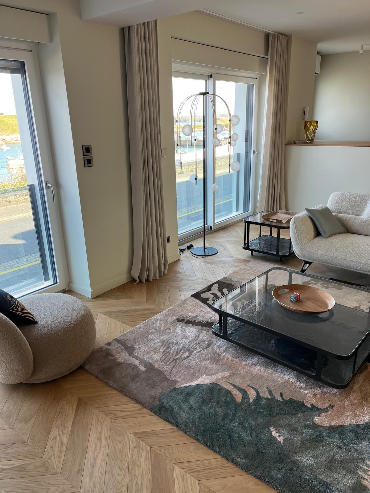 Salon lumineux avec portes-fenêtres, canapé beige, tapis à motifs et table basse en verre.