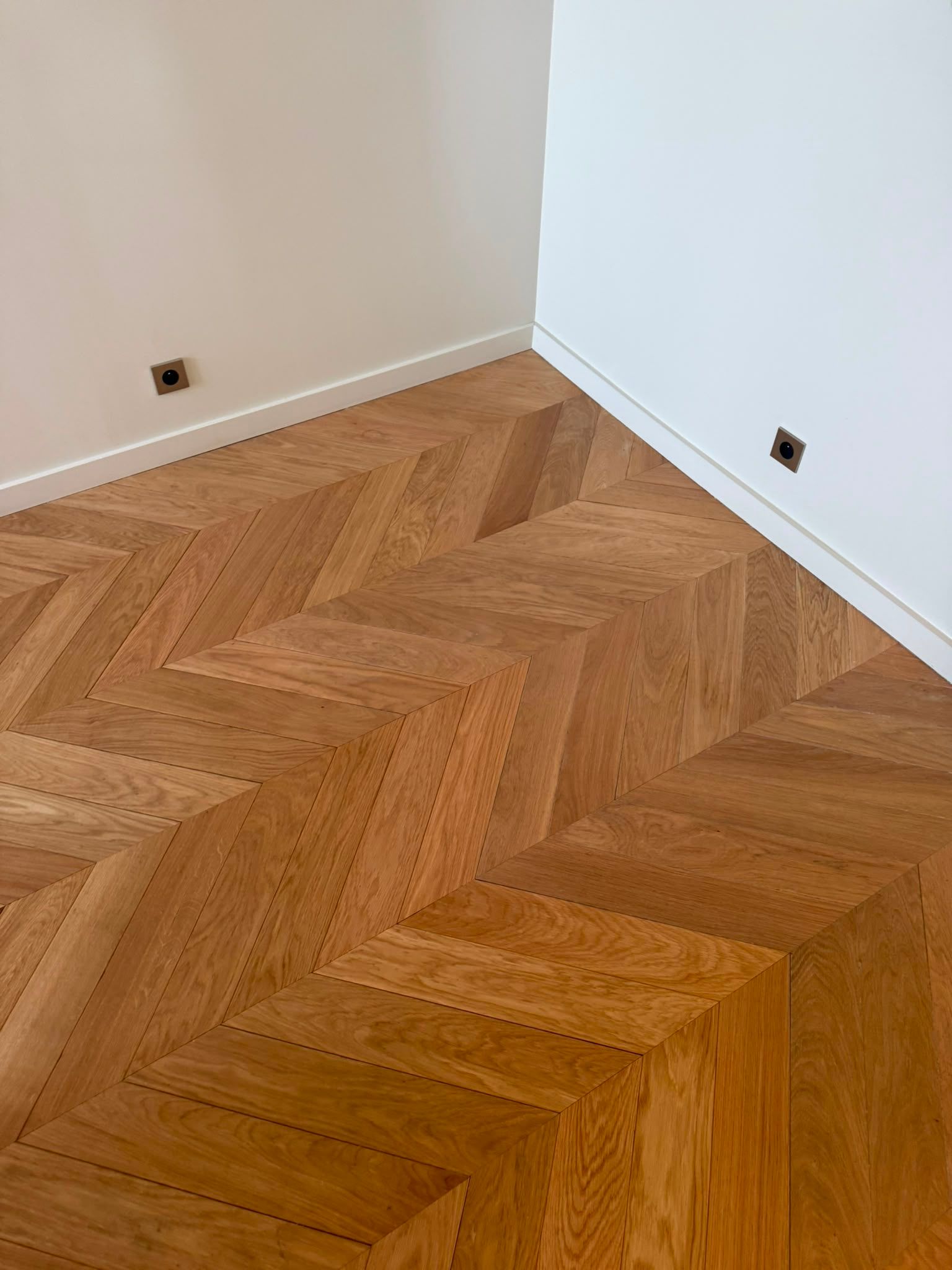 Coin d’une pièce avec un parquet en chevrons, des murs blancs et des prises électriques.