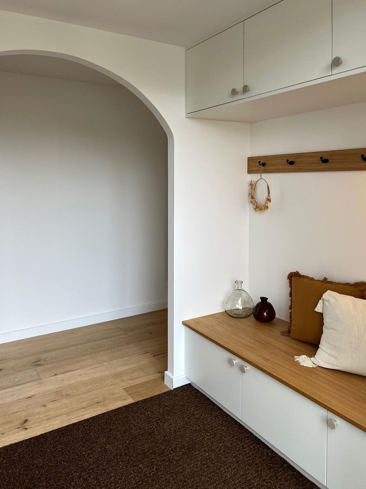 Coin de chambre minimaliste avec rangements intégrés blancs, banc en bois, tapis marron et porte cintrée.