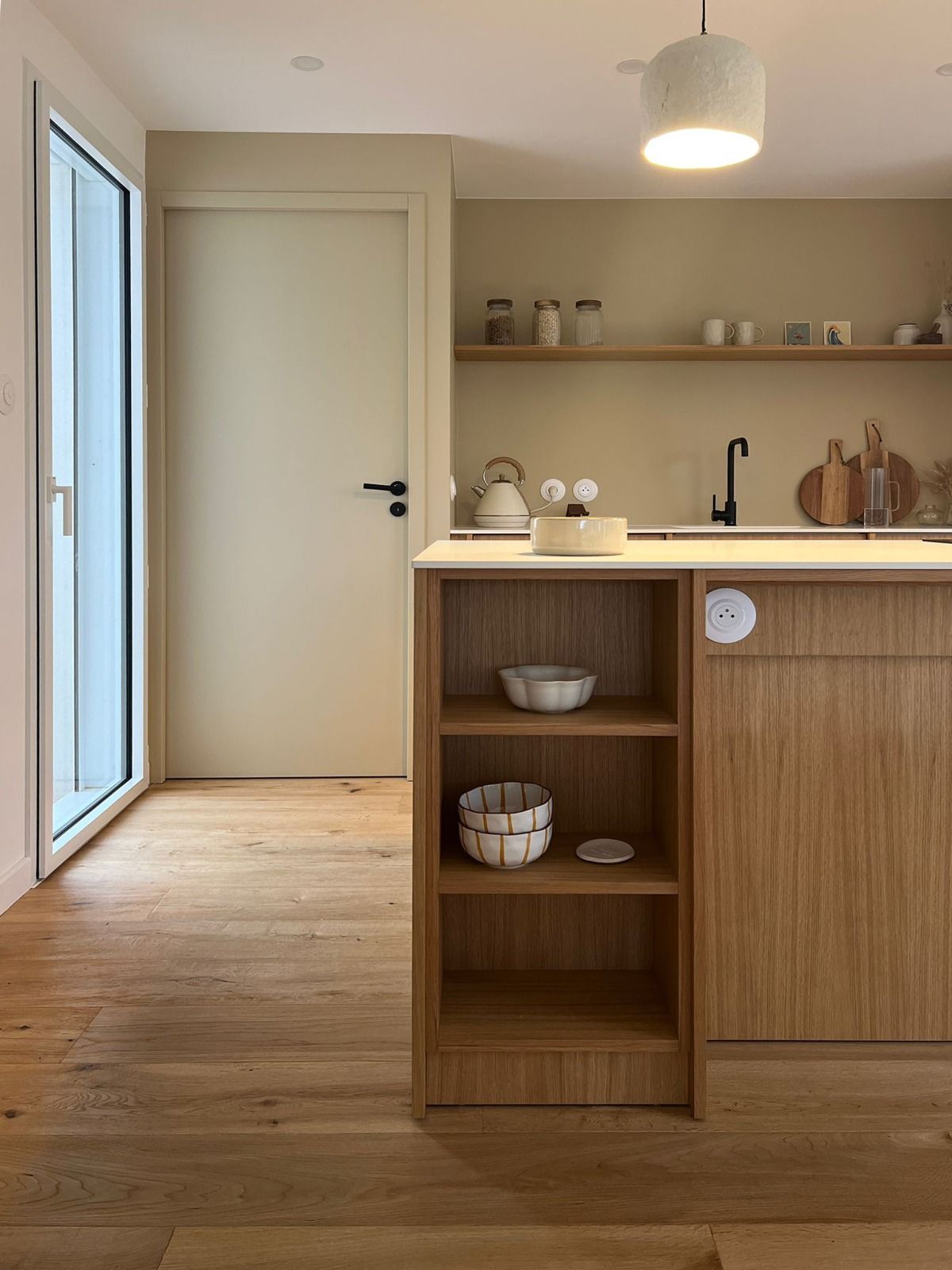 Cuisine lumineuse et minimaliste avec des placards en bois, des étagères ouvertes, un évier et une porte vitrée à gauche.