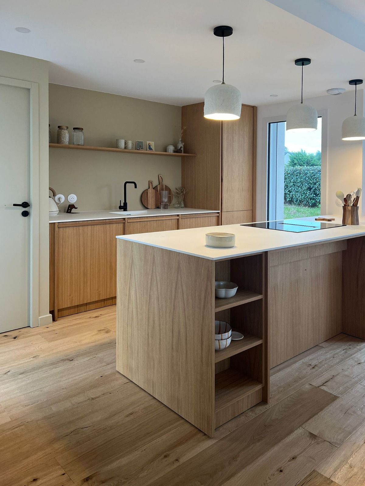 Cuisine moderne avec îlot en bois, étagères ouvertes, suspensions et grandes fenêtres donnant sur la verdure