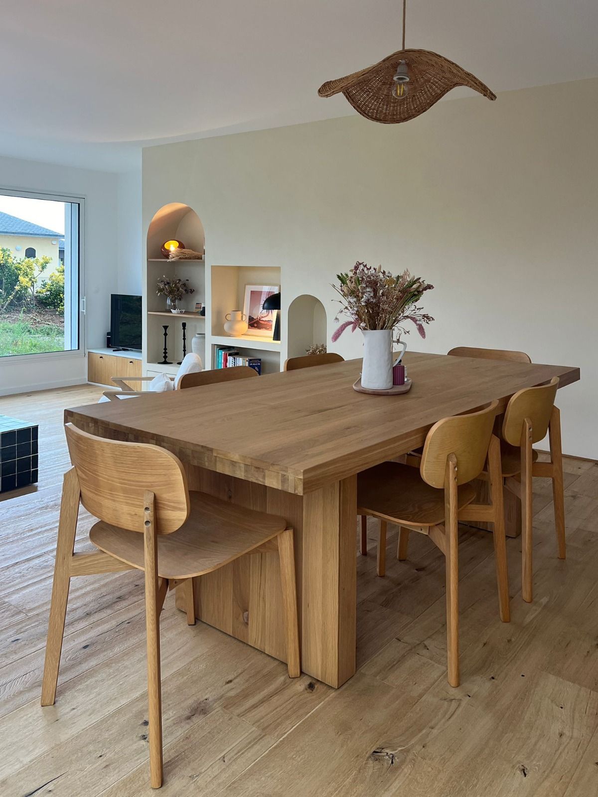 Salle à manger lumineuse avec une grande table en bois, quatre chaises et un vase de fleurs séchées près d’une fenêtre.