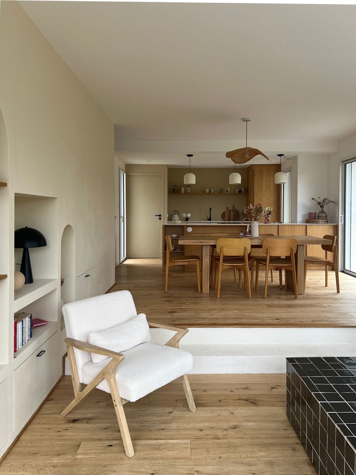 Salon lumineux avec un fauteuil blanc, un parquet, une table à manger et une cuisine ouverte en arrière-plan