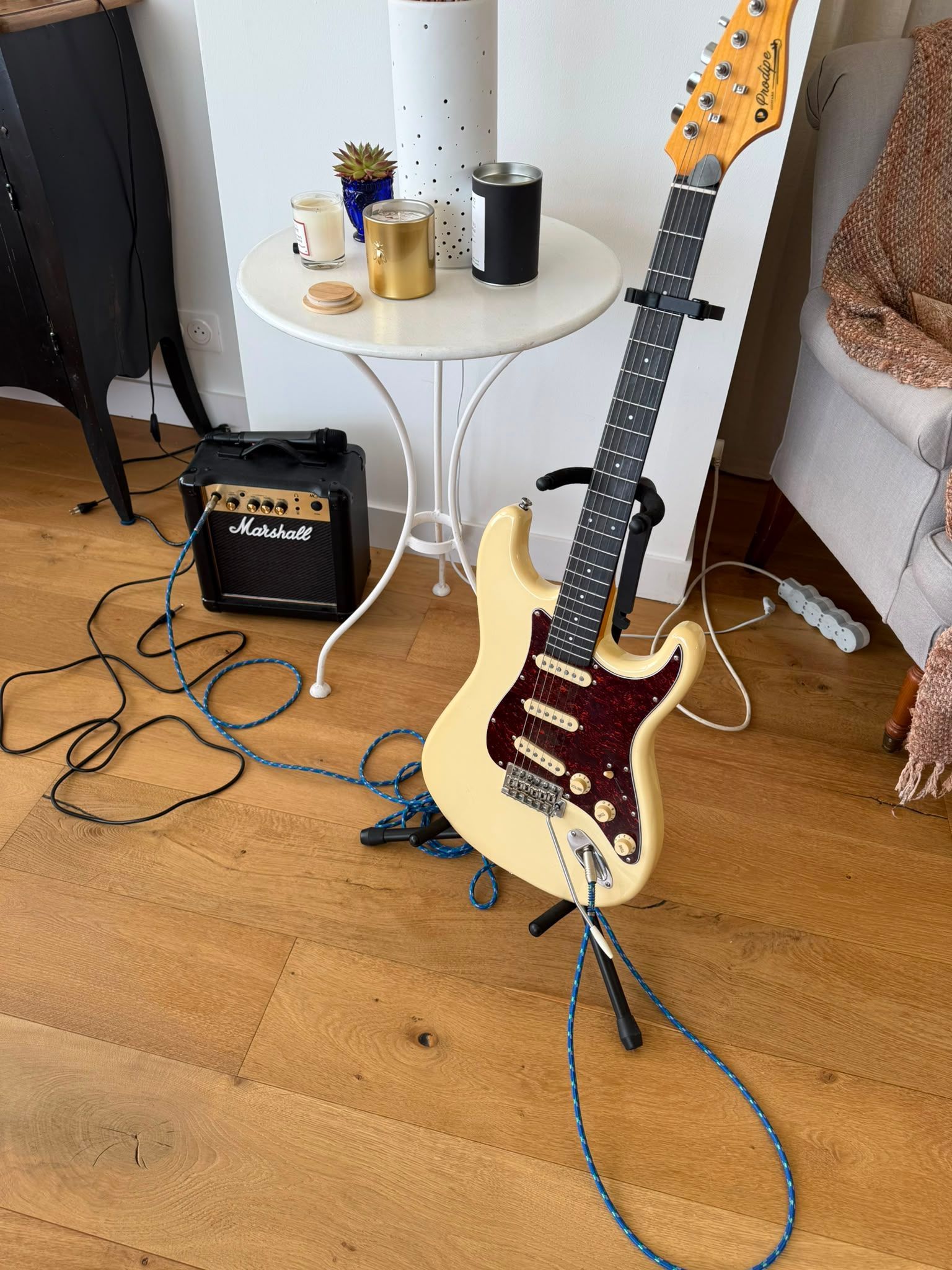 Guitare électrique sur un support, à côté d’une petite table avec des mugs, des câbles et un amplificateur, sur un parquet.