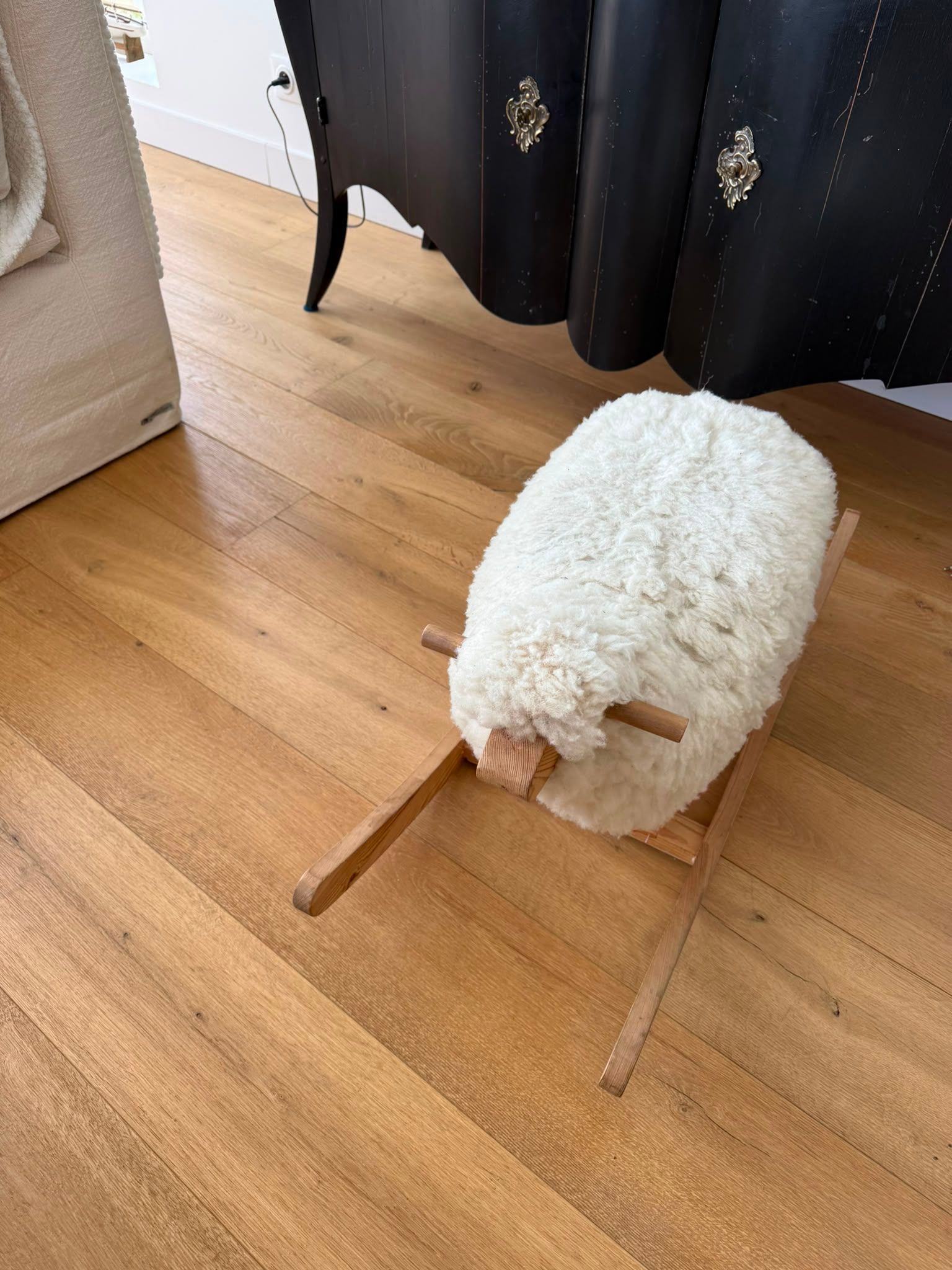 Petit tabouret blanc duveteux ou panier pour animal de compagnie posé sur un parquet, près d’un rideau et de meubles.