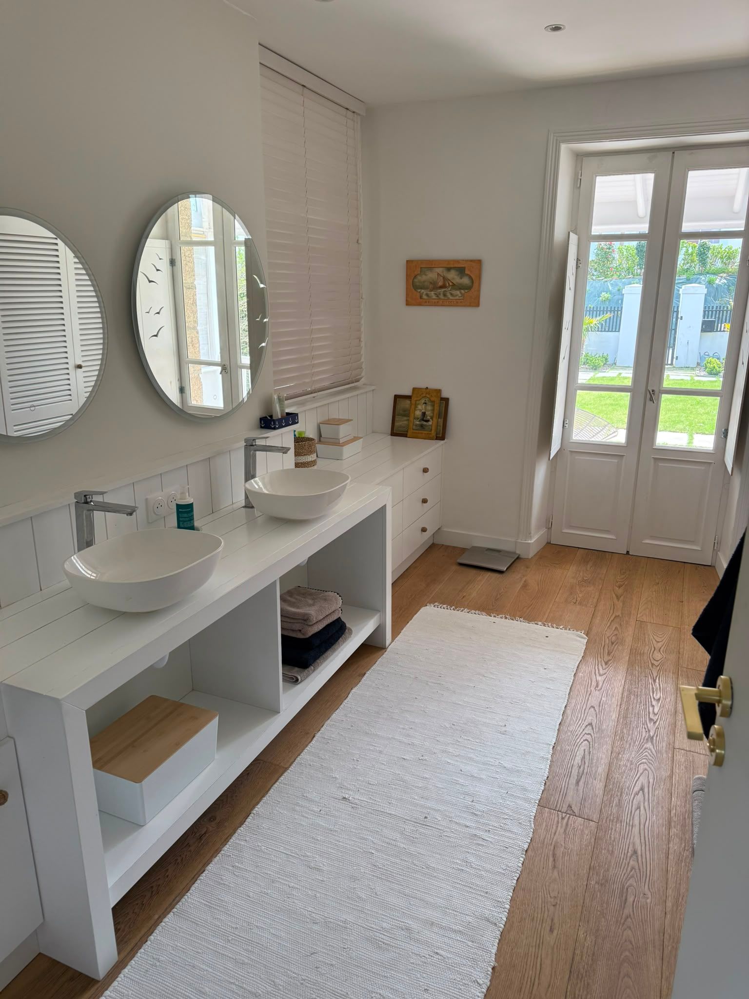 Salle de bains lumineuse avec deux lavabos, des miroirs ronds, un tapis blanc et des portes-fenêtres donnant sur un jardin.