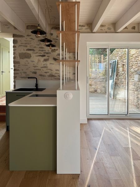 Cuisine moderne et coin repas avec îlot vert, poutres en bois, mur en pierre et portes coulissantes en verre