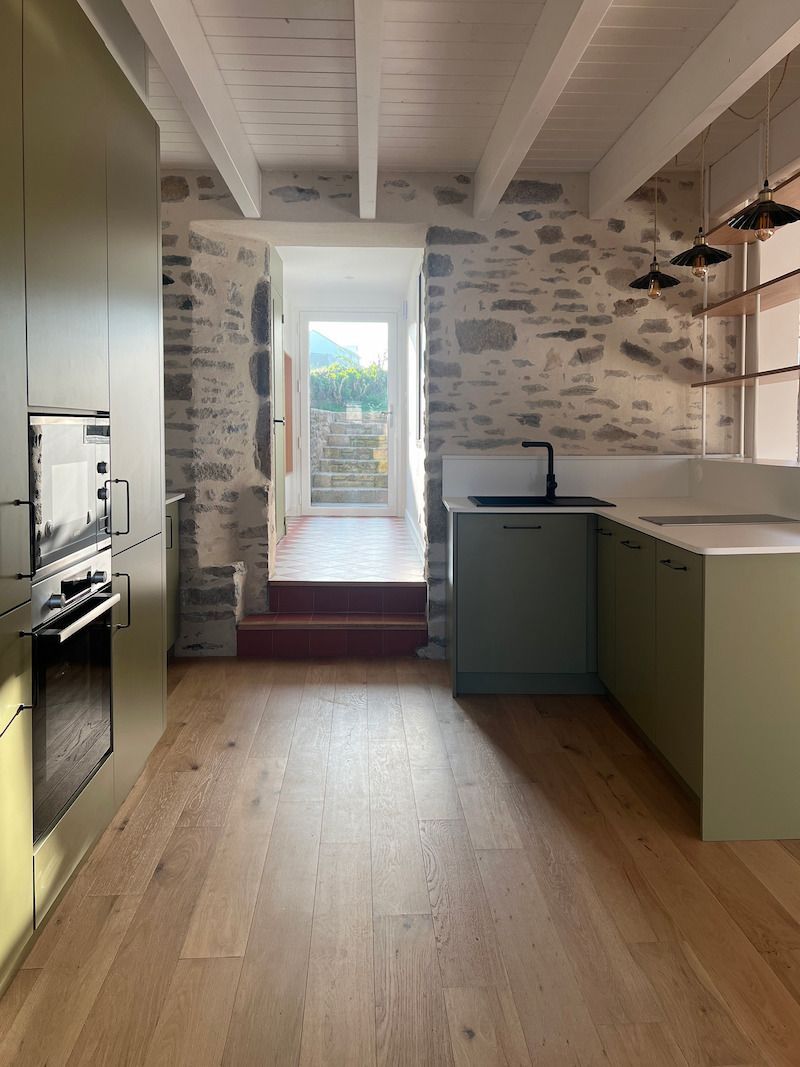 Cuisine rustique et étroite, avec un parquet, un mur en pierre, des placards verts et une porte lumineuse au fond