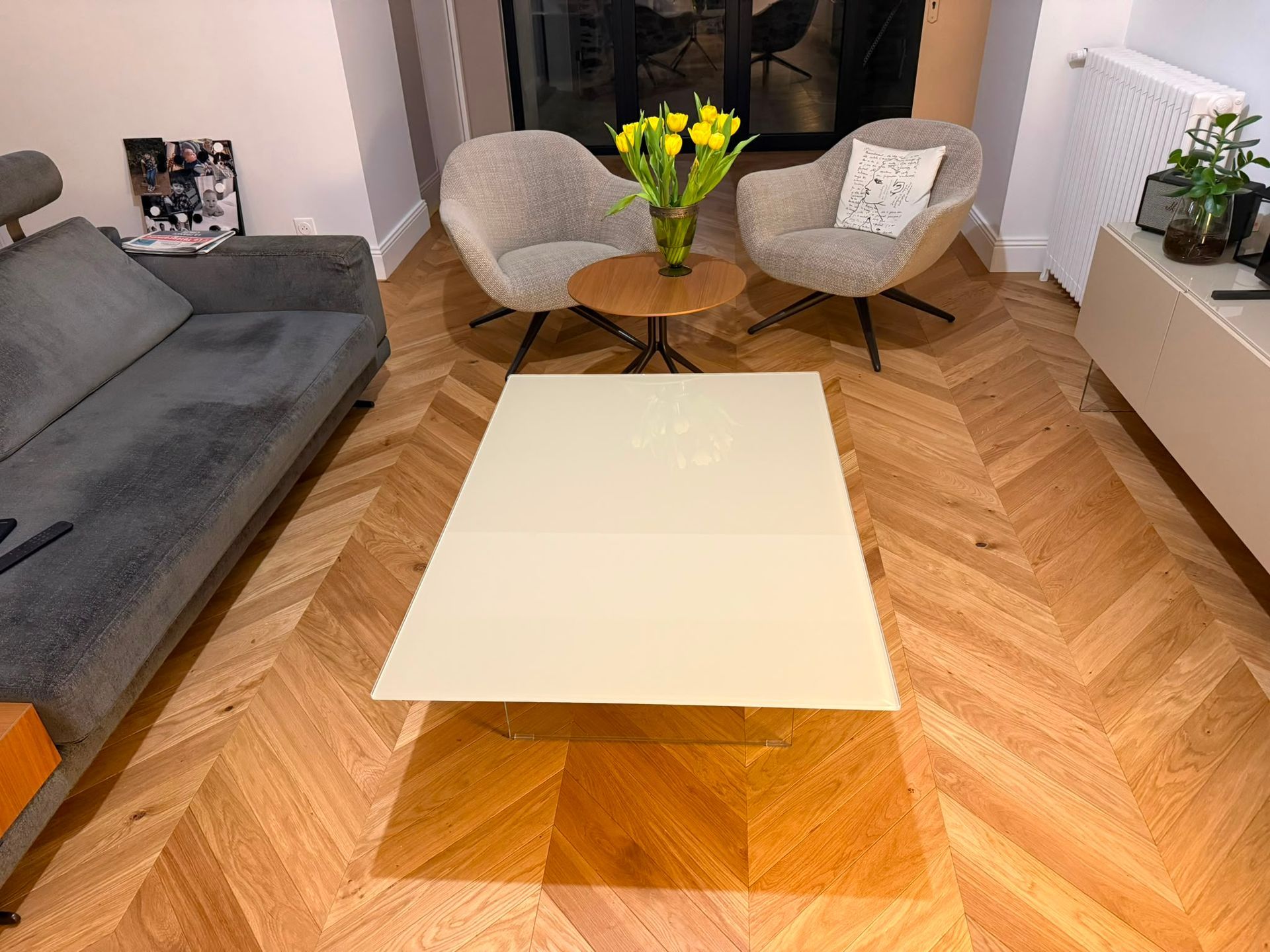 Salon moderne avec un canapé gris, une table basse blanche, deux fauteuils et un vase de fleurs jaunes