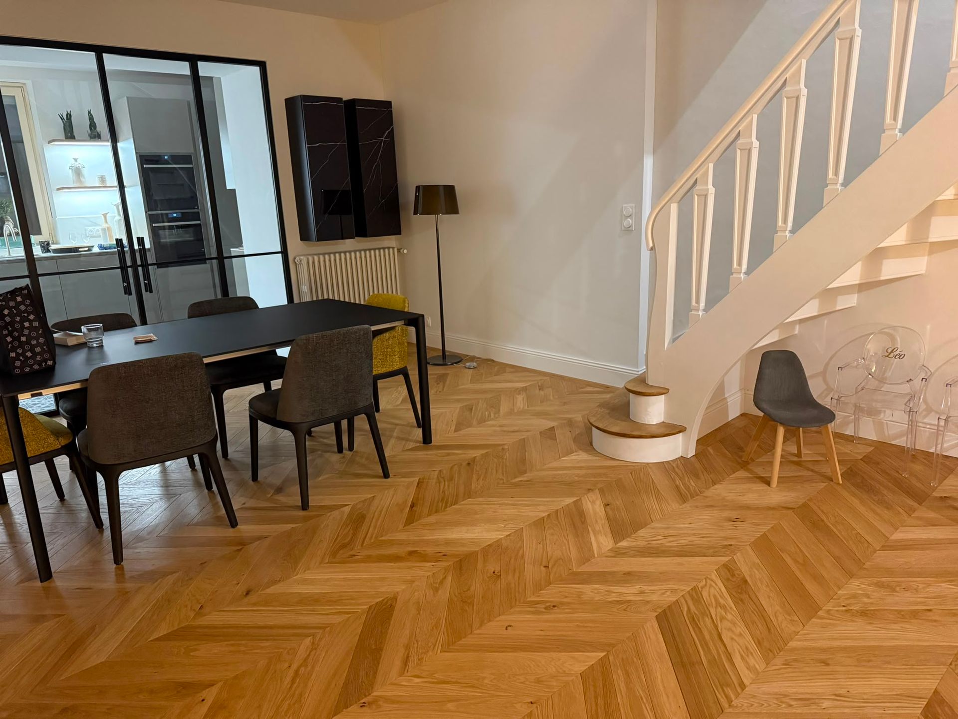 Salle à manger avec parquet en chevrons, table et chaises, et escalier à droite.