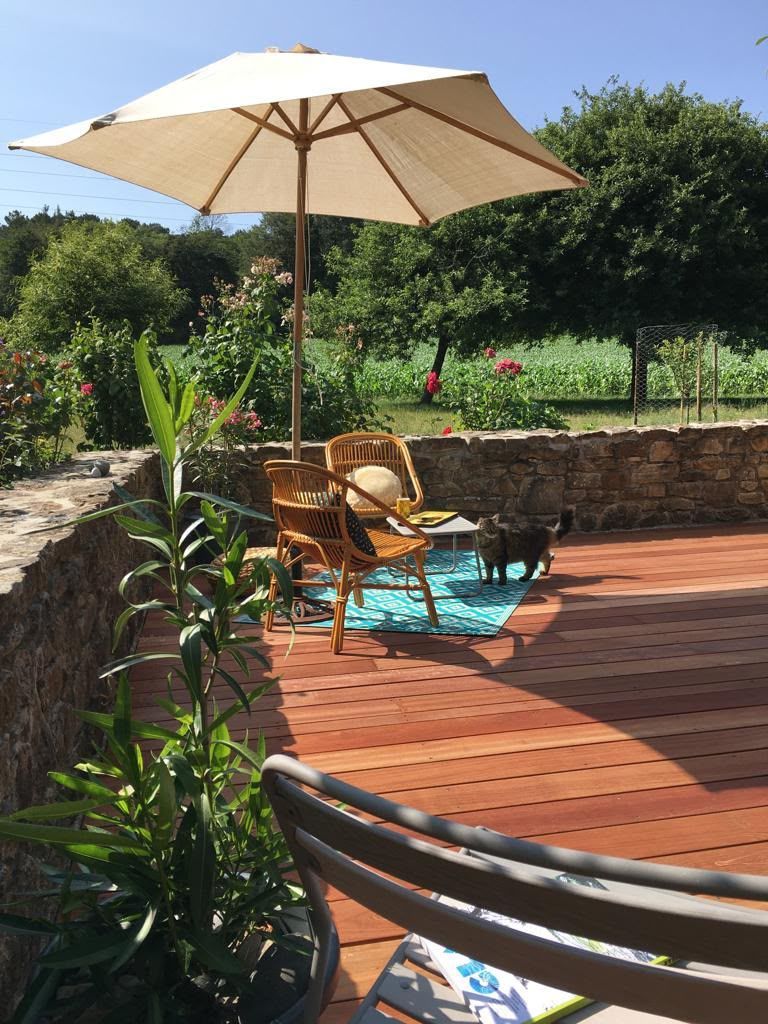 Terrasse avec des chaises longues en bois sous un parasol crème, à côté d’un mur en pierre et d’un jardin verdoyant.