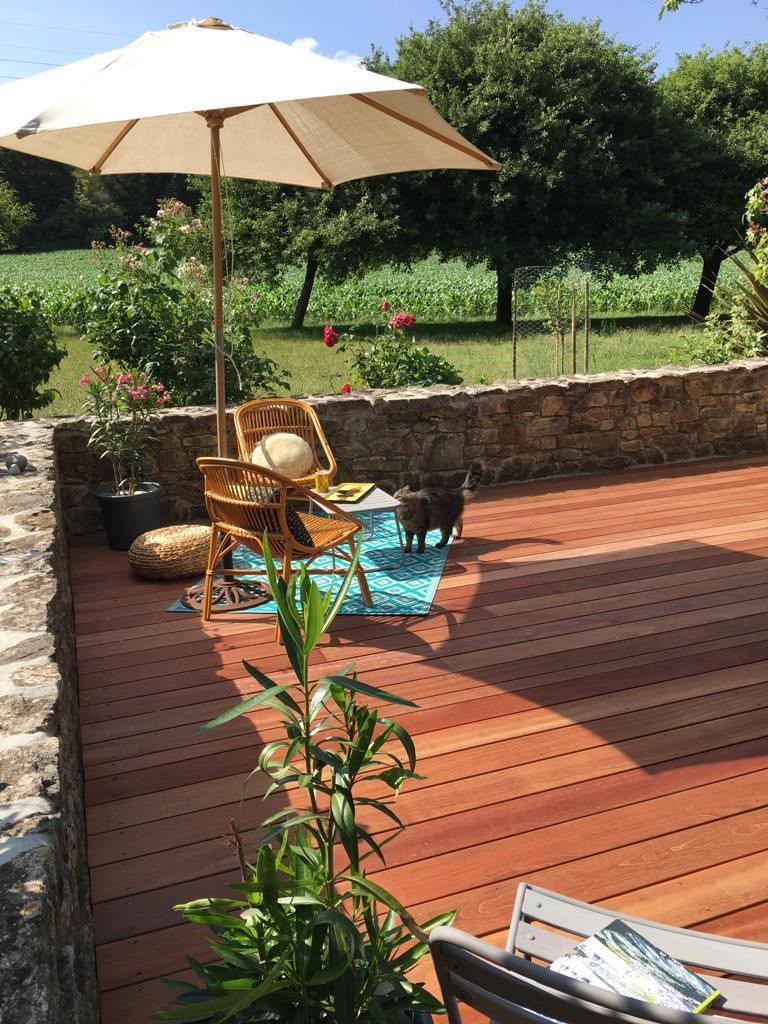 Patio avec terrasse en bois, parasol blanc, fauteuil en osier et plantes en pot, près d’un mur de pierre et d’un jardin.