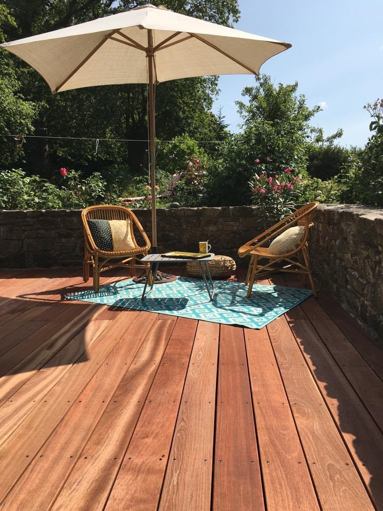 Terrasse avec deux chaises, un parasol blanc et une petite table posés sur un tapis turquoise dans un jardin ensoleillé.