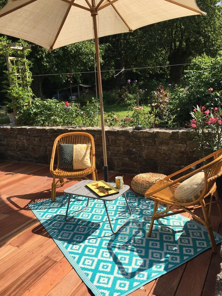 Coin de terrasse avec deux fauteuils en osier, une petite table et un tapis turquoise sous un parasol beige ensoleillé