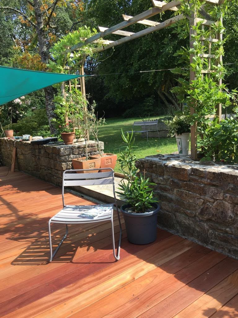 Patio avec une terrasse en bois, une chaise, une plante en pot, des murs en pierre et un jardin verdoyant au-delà