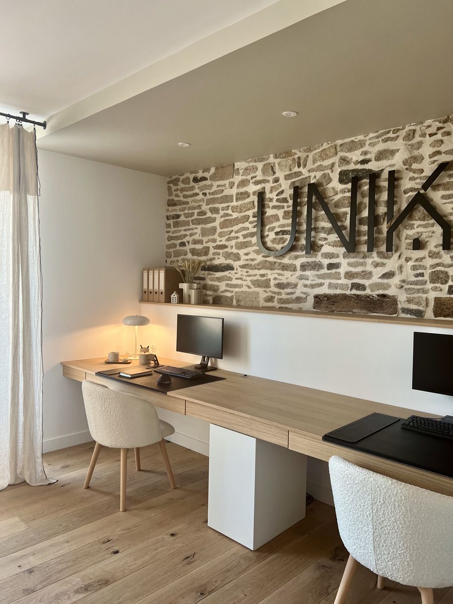 Bureau à domicile moderne avec deux bureaux, des écrans, des chaises et un mur en pierre orné du lettrage « UNIKA ».