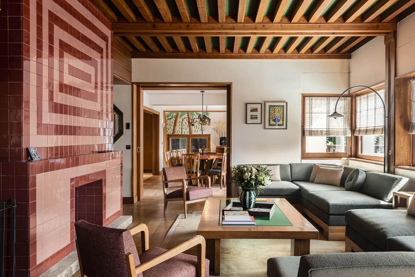 Salon confortable avec une cheminée en briques rouges, un canapé d’angle gris, des poutres en bois et de grandes fenêtres