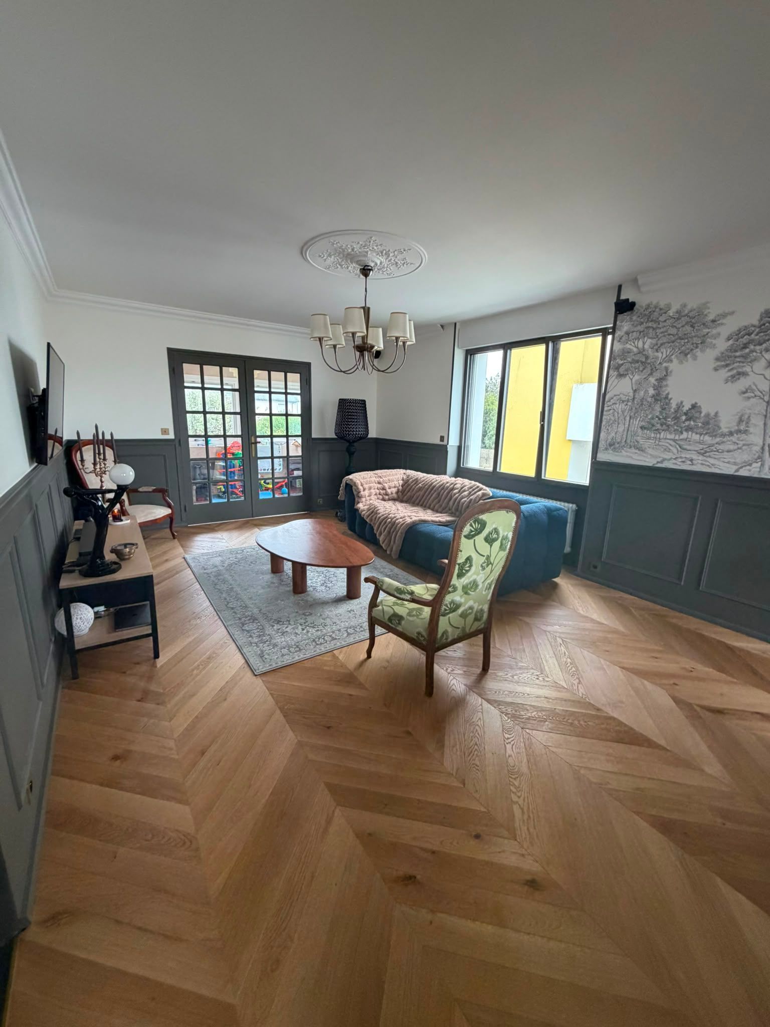 Salon lumineux avec parquet en chevrons, canapé à motifs, téléviseur, lustre et grandes fenêtres.
