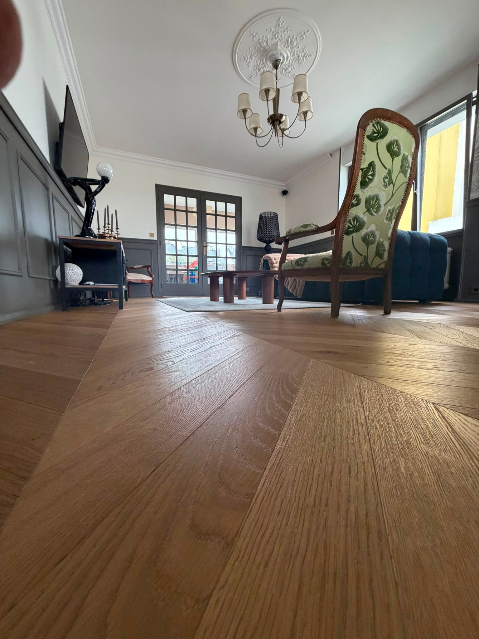 Vue en contre-plongée d’un salon lumineux avec parquet, lustre, fauteuils et cheminée.