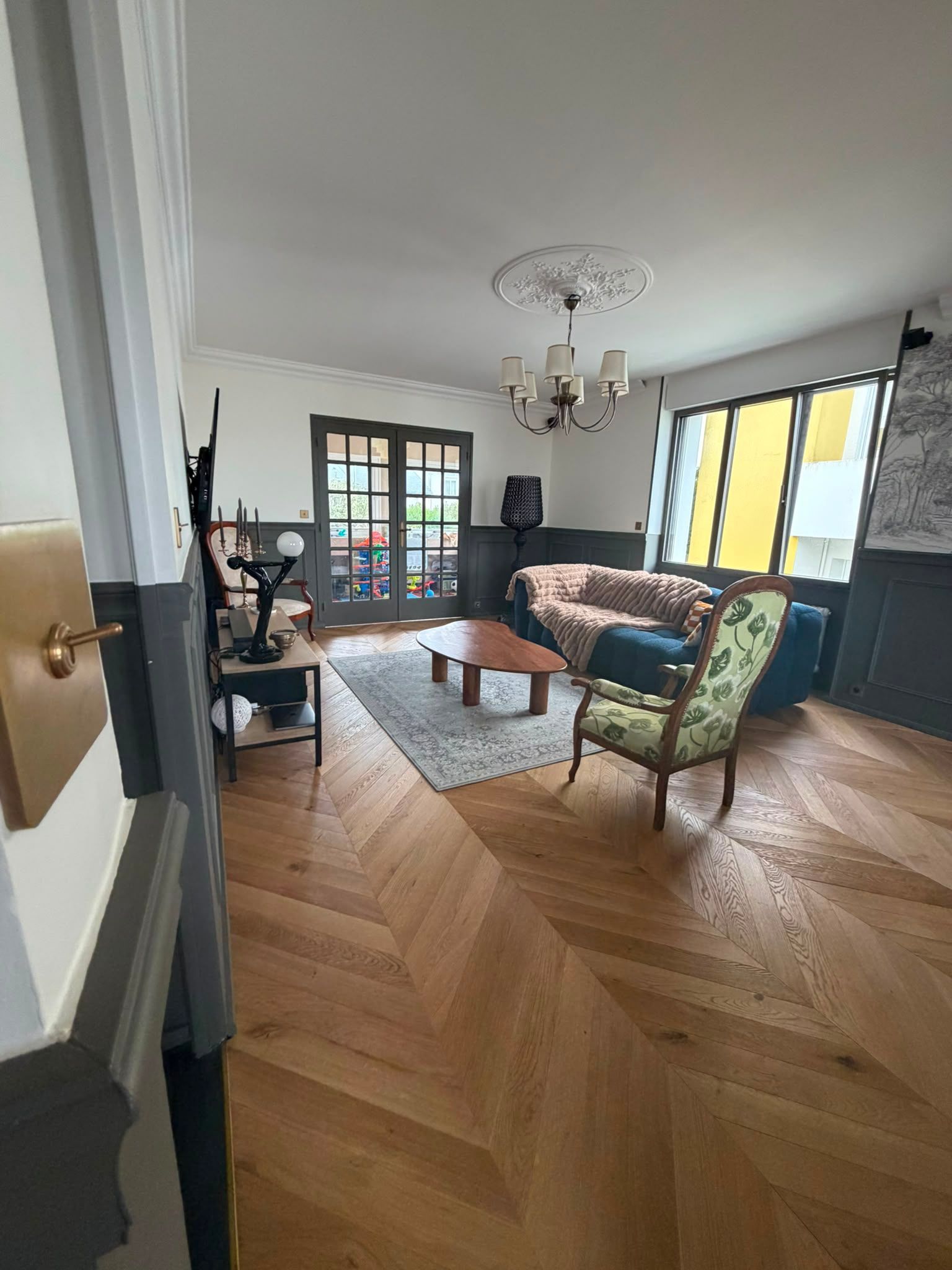 Salon lumineux avec parquet en chevrons, canapé à motifs, table basse en verre et grandes fenêtres.